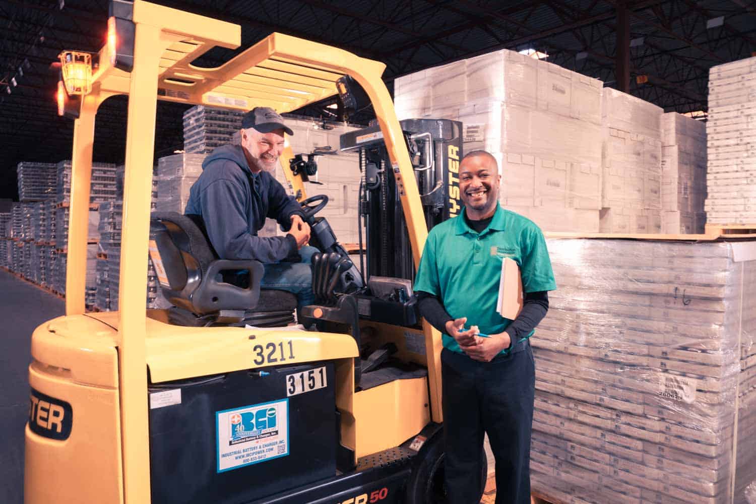 person in a forklift and another person standing outside the forklift, both smiling at camera; background is pallets