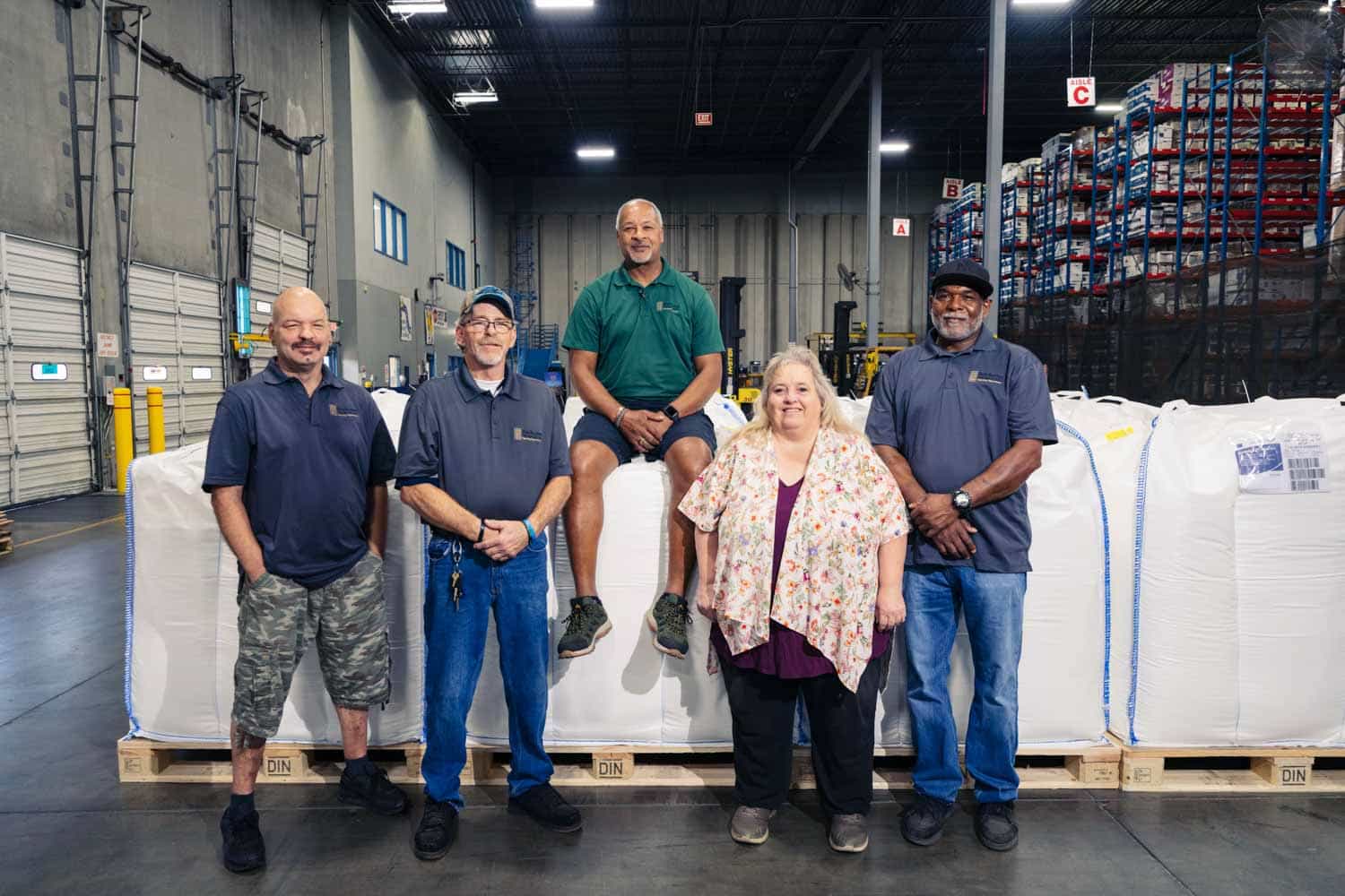 A Team, representing Distribution Technology value of teamwork; 5 individuals standing and sitting on pallets inside a warehouse