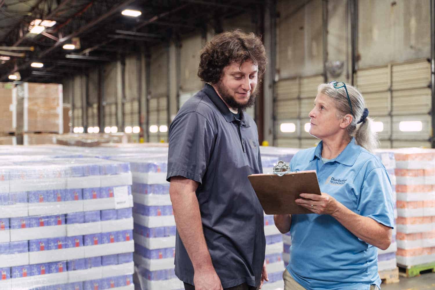 Distribution Technology value - Quality; Tall young man talk with a shorter lady with clipboard in her hand; background is warehouse