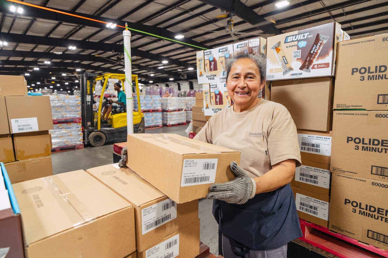 mature woman with gloves on holding a box and smiling at the camera; background is interior warehouse