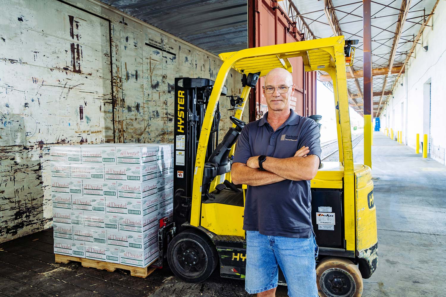 forklift worker standing with arms folded in front of yellow forklift with full load of pallets